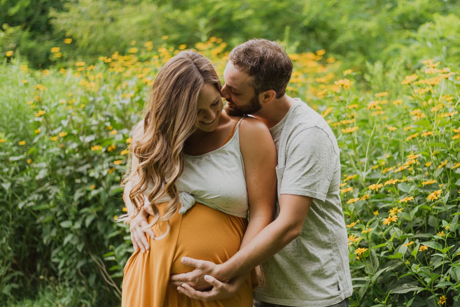 Pregnant couple embracing in a field of yellow wildflowers, man kissing woman's head during maternity photo session.
