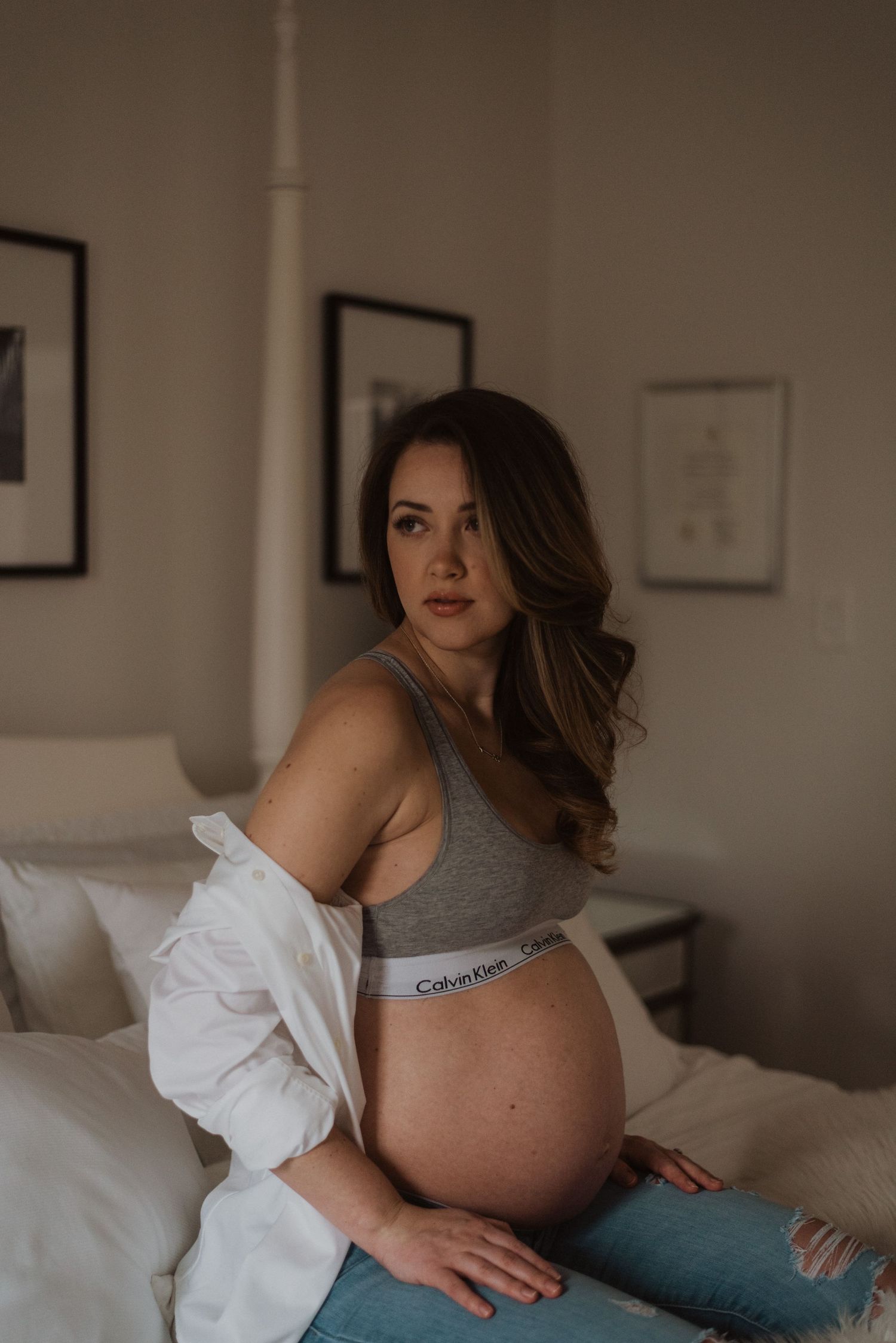Pregnant woman in Calvin Klein underwear and open white shirt sitting on a bed for a maternity photoshoot.