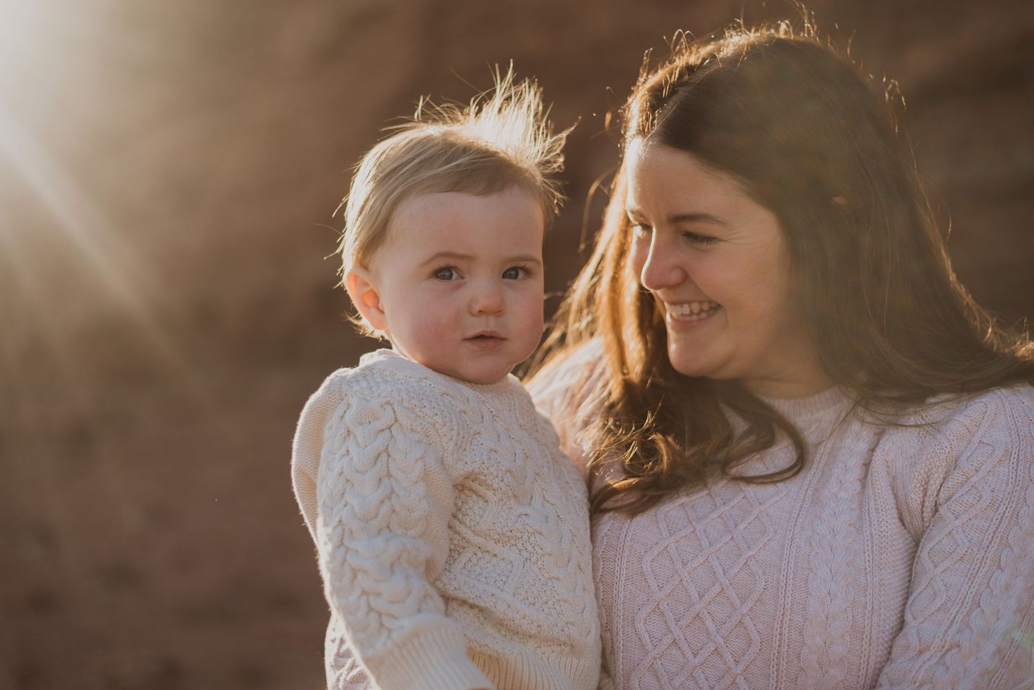 A mother smiles warmly at her baby in golden sunlight, both wearing cream knit sweaters outdoors.