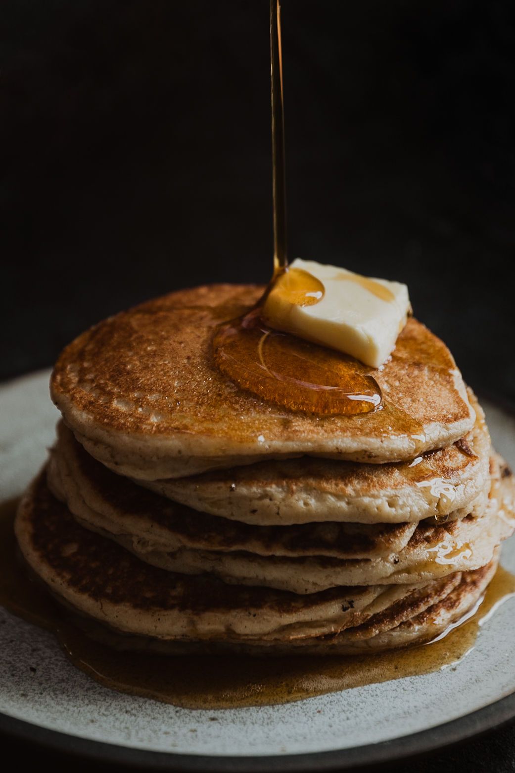 Stack of fluffy pancakes topped with butter and maple syrup drizzling down on a dark background.
