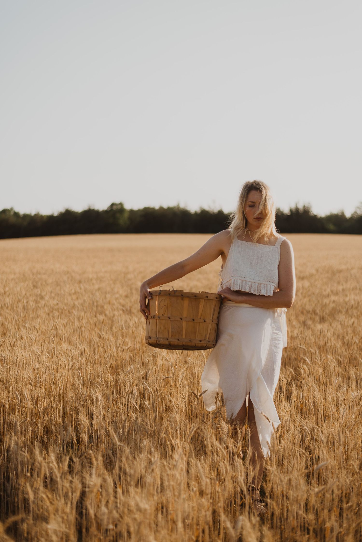 Blonde woman in white dress holding a wicker basket standing in a golden wheat field at sunset.