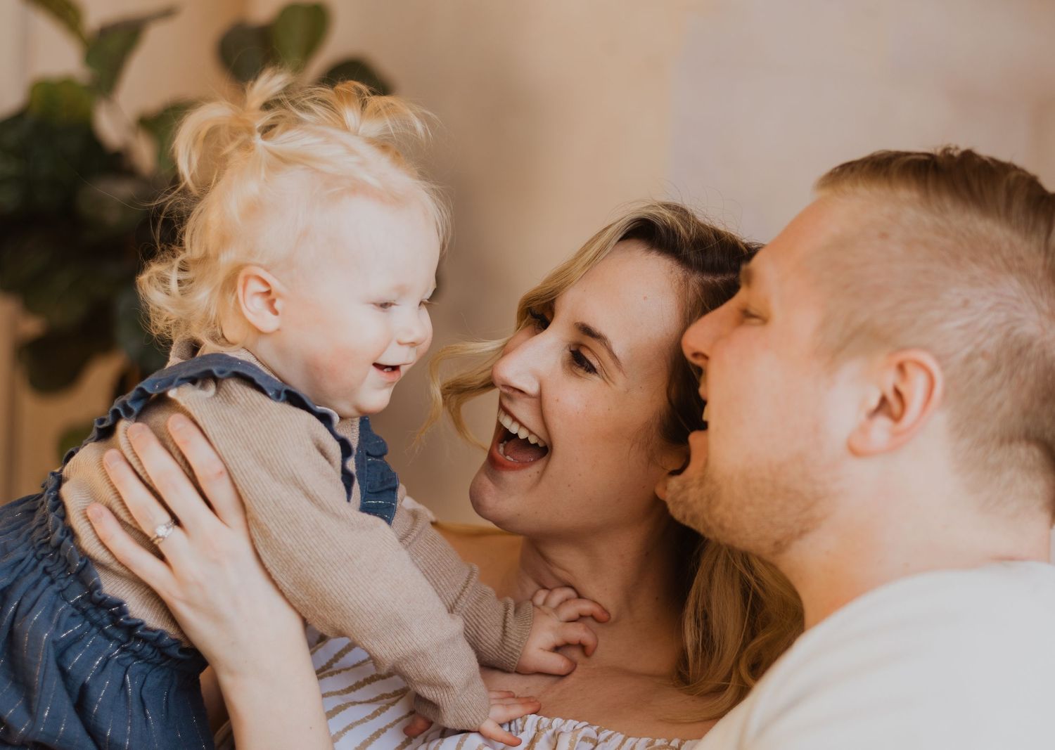 Happy family moment with laughing mother holding blonde toddler girl while smiling father looks on warmly.