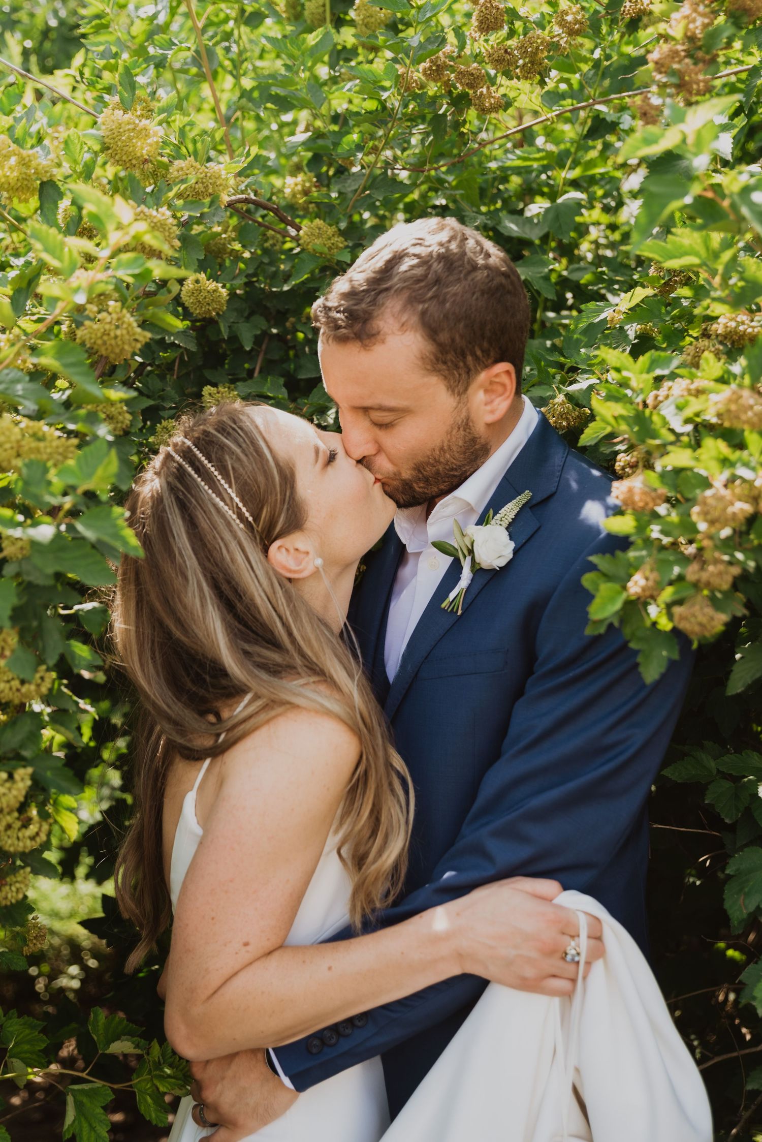 Bride and groom sharing a romantic kiss in front of lush green foliage, bride wearing white gown and groom in navy suit.