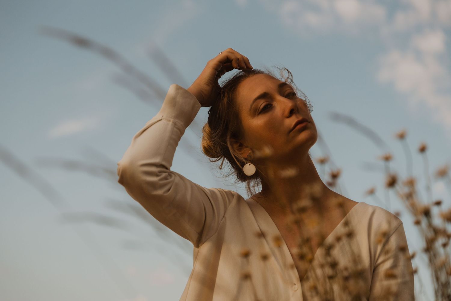 Woman in white cardigan poses outdoors against blue sky, hand in hair, with dried wildflowers in golden light.