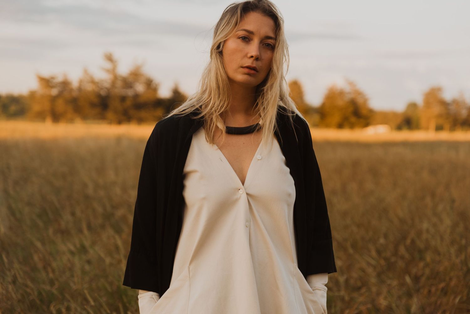 Blonde woman in white slip dress and black cardigan standing in golden field at sunset.