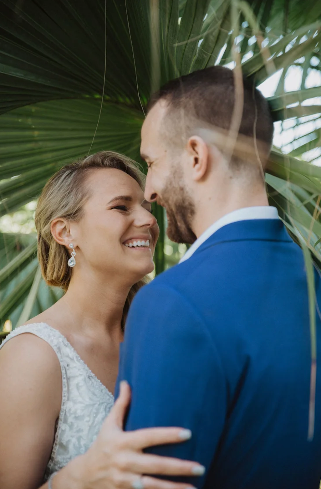 A smiling bride and groom share a romantic moment under tropical palm leaves, with the groom in a blue suit.