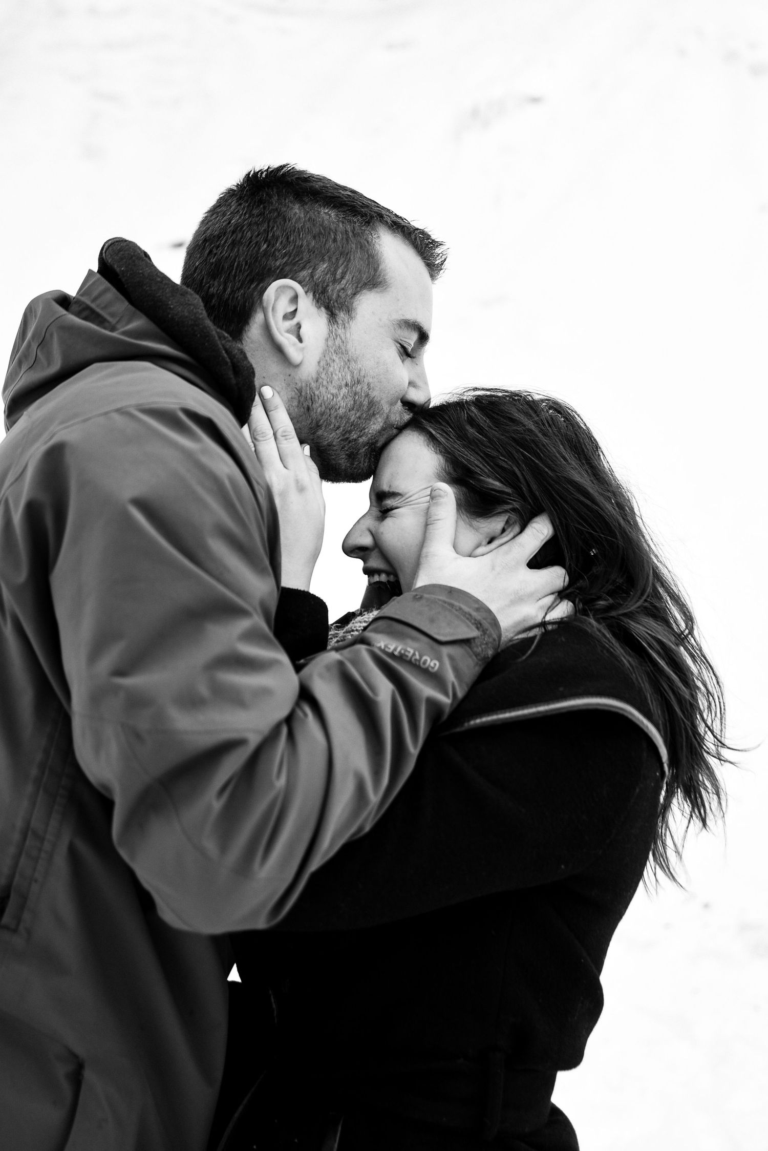 Black and white photo of a couple sharing a tender forehead kiss outdoors in winter, man gently holding womans face.