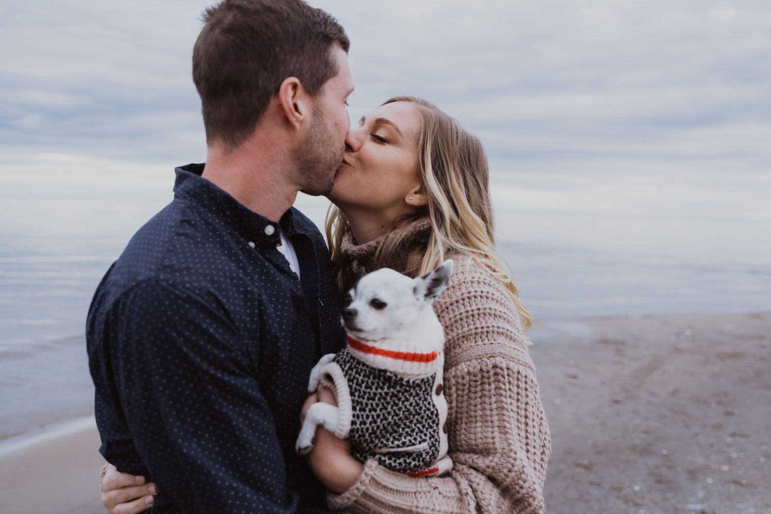 Couple kissing on a beach while holding a small white dog in a sweater.