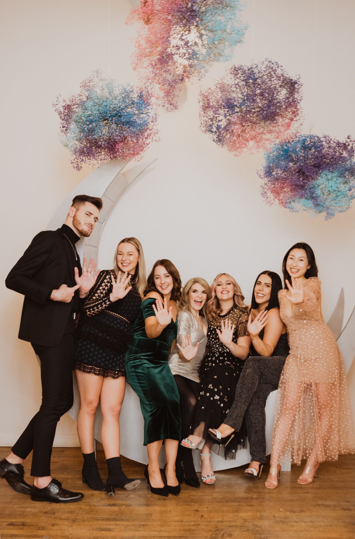 Group of seven friends posing in elegant party attire in front of colorful floral wall art installation.