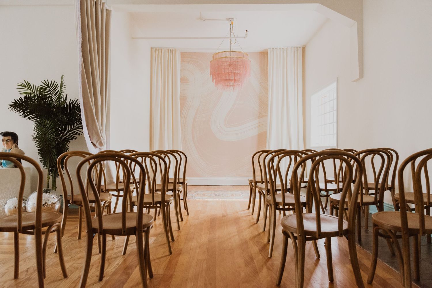 Elegant wedding ceremony setup with bentwood chairs, sheer curtains, a pink chandelier, and a blush floral backdrop on hardwood floors.