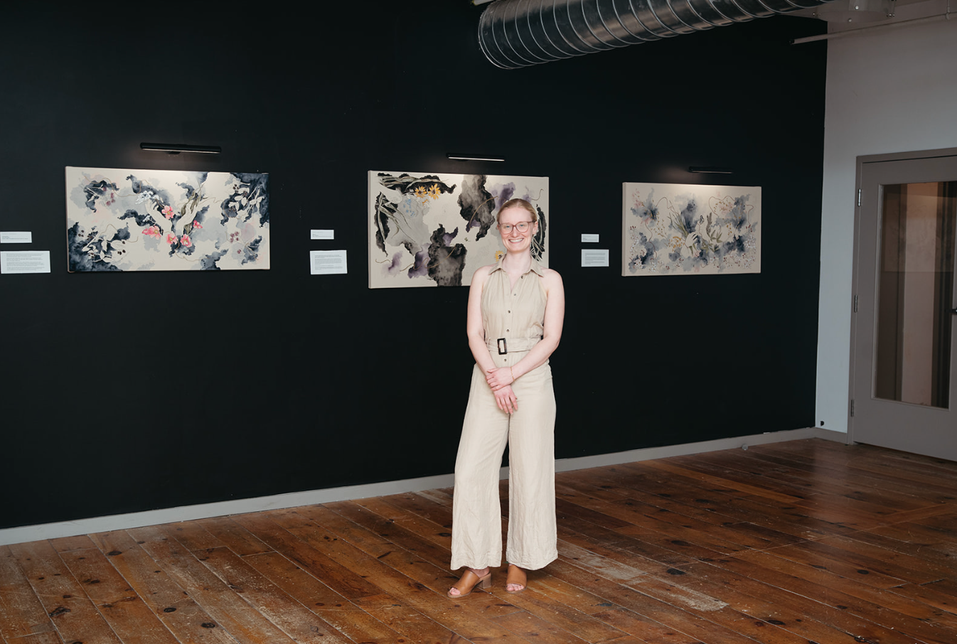Woman standing in art gallery with abstract paintings on dark walls and hardwood floors.