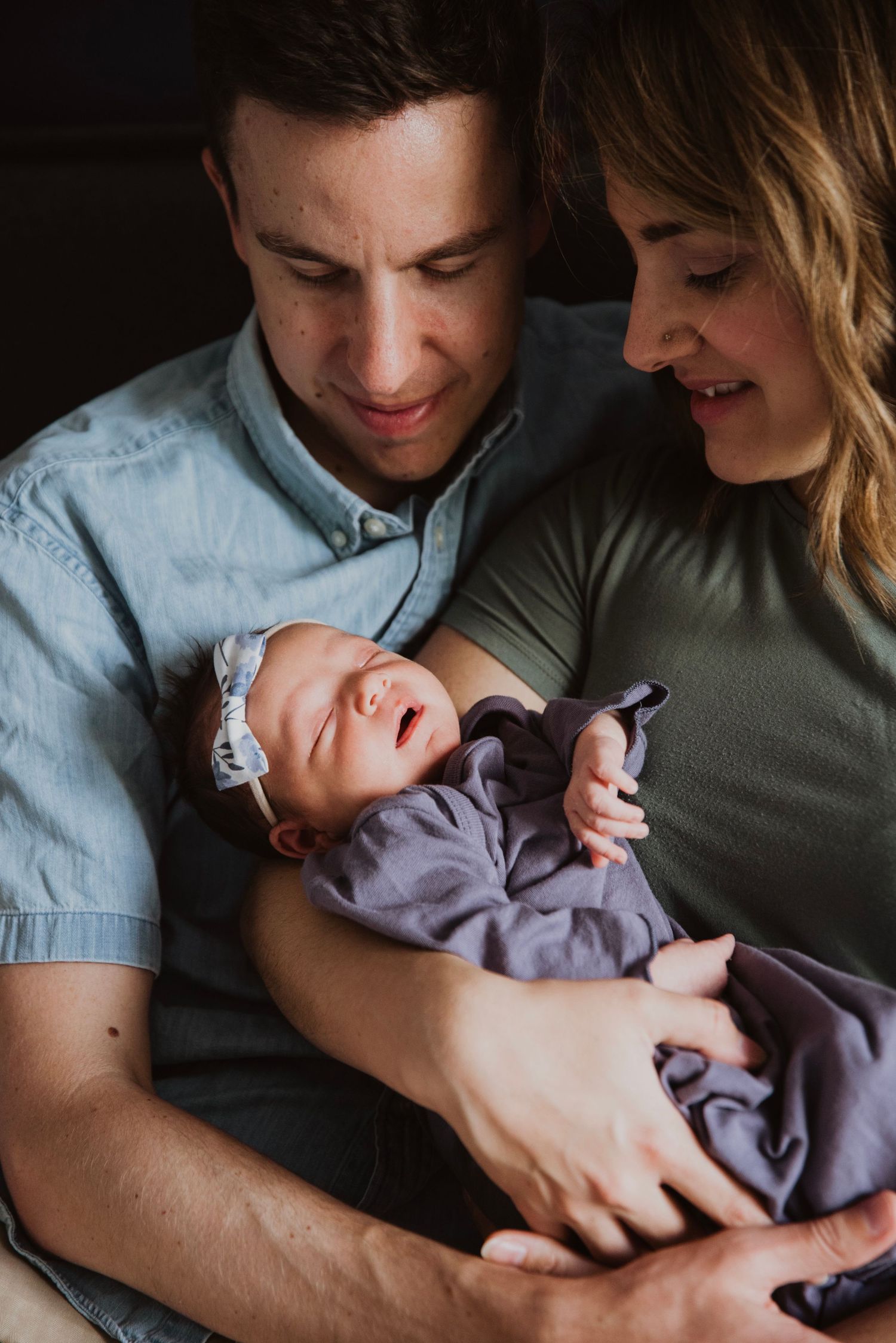 New parents smiling lovingly at their newborn baby wrapped in purple, with mom in green and dad in blue shirt.