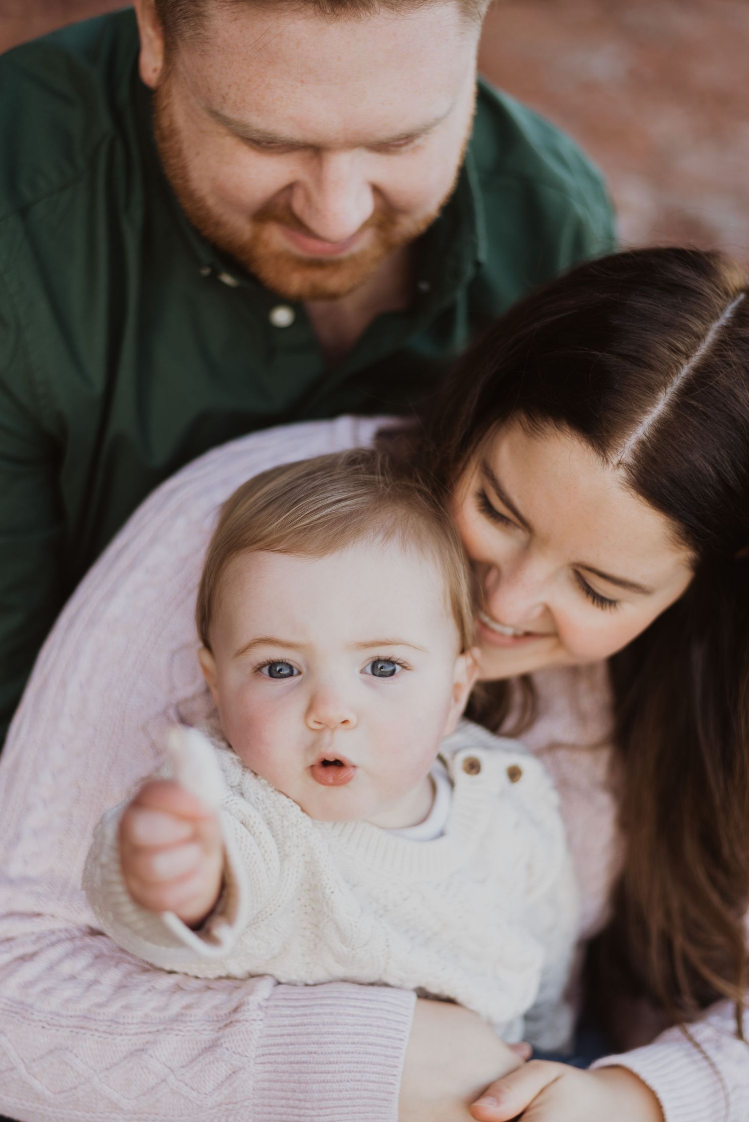 A happy young family with a bearded father, smiling mother, and curious blue-eyed baby in a white knit sweater.