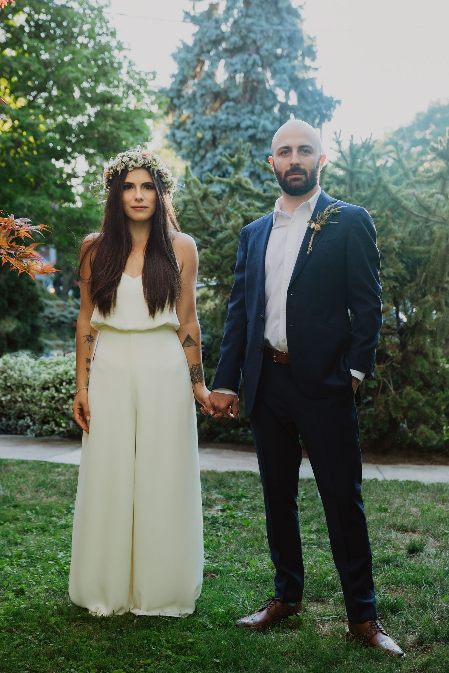 Bride in white jumpsuit with floral crown and groom in navy suit holding hands outdoors at wedding.