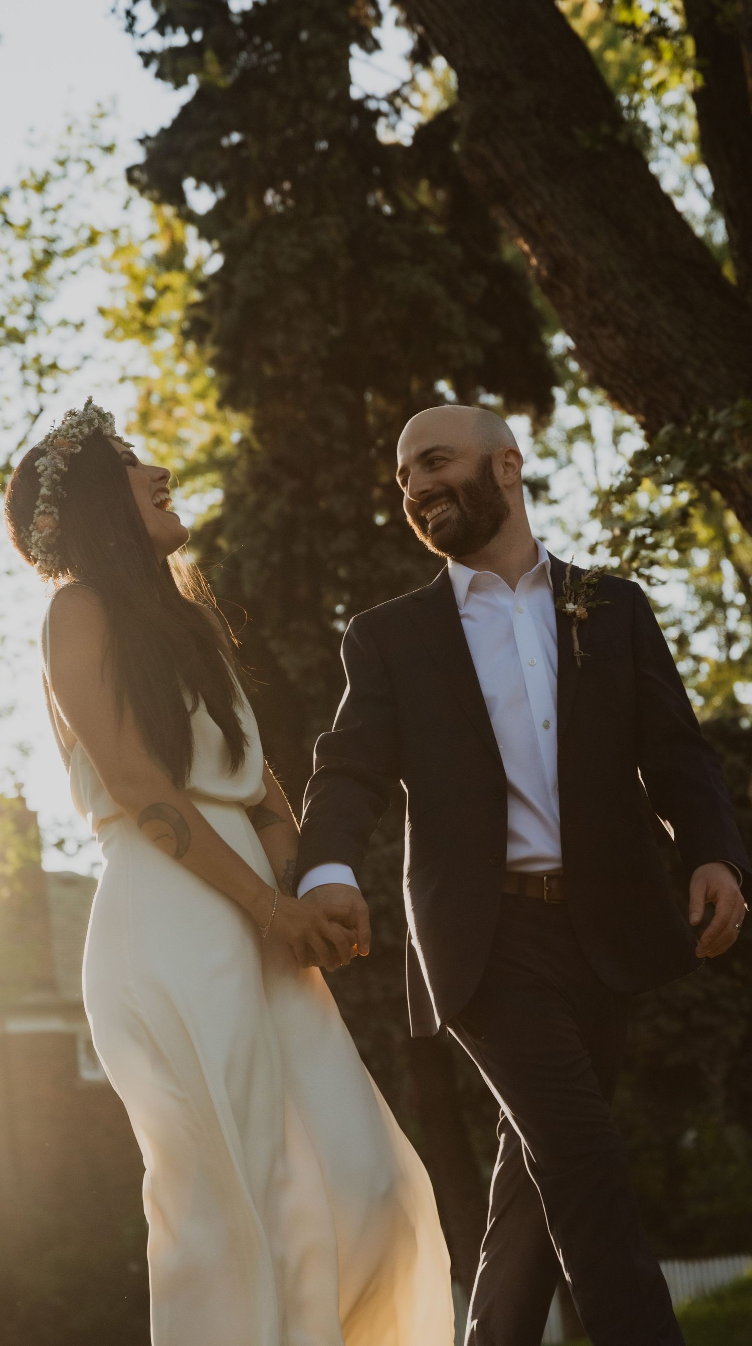 A joyful bride and groom hold hands and laugh together outdoors in golden sunset light near a large tree.