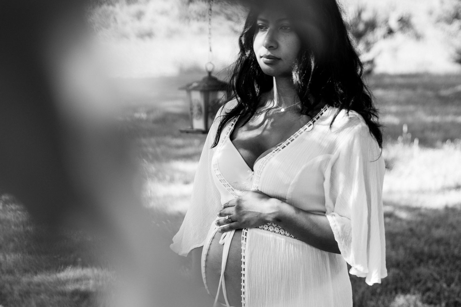 Black and white photo of a pregnant woman in a white dress cradling her baby bump outdoors.