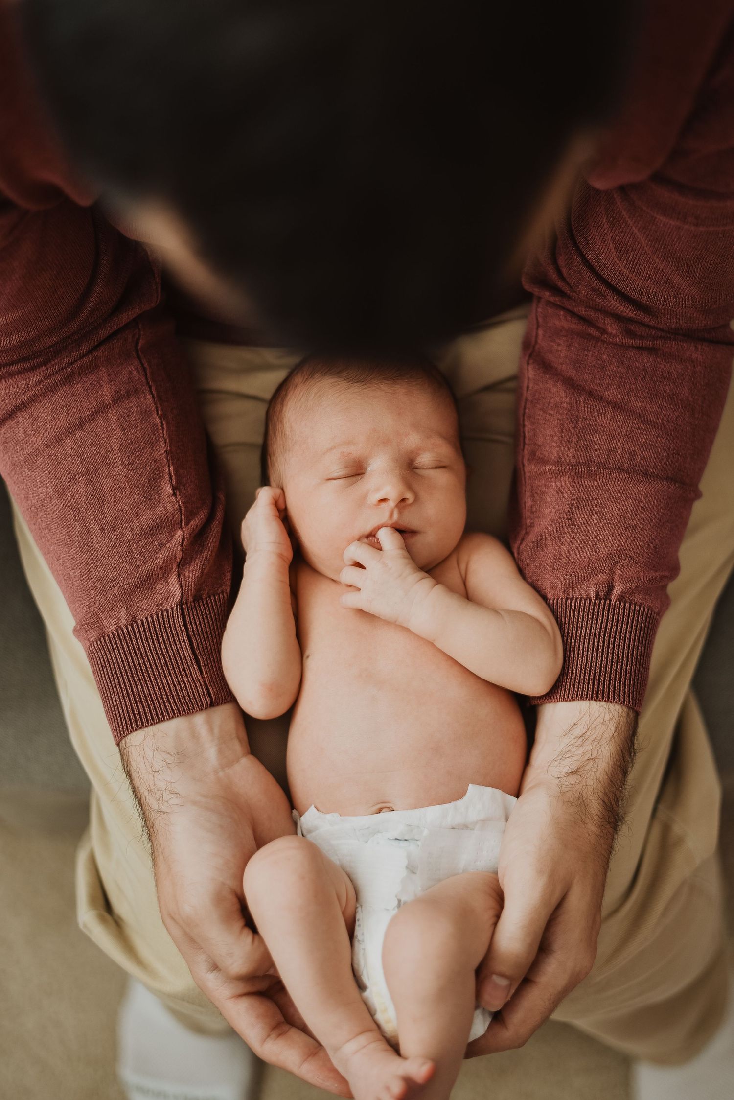Newborn baby sleeping peacefully in father's hands, wearing a diaper, with dad in a red sweater.