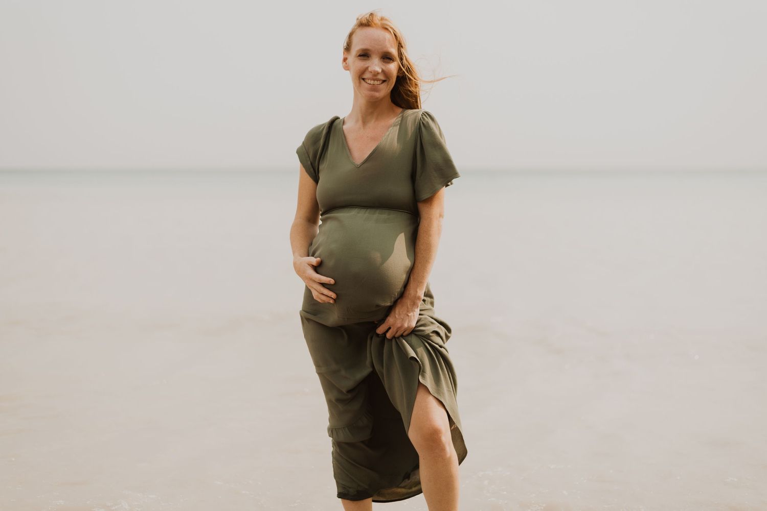 Smiling pregnant woman in olive green wrap dress cradling baby bump on a serene beach.