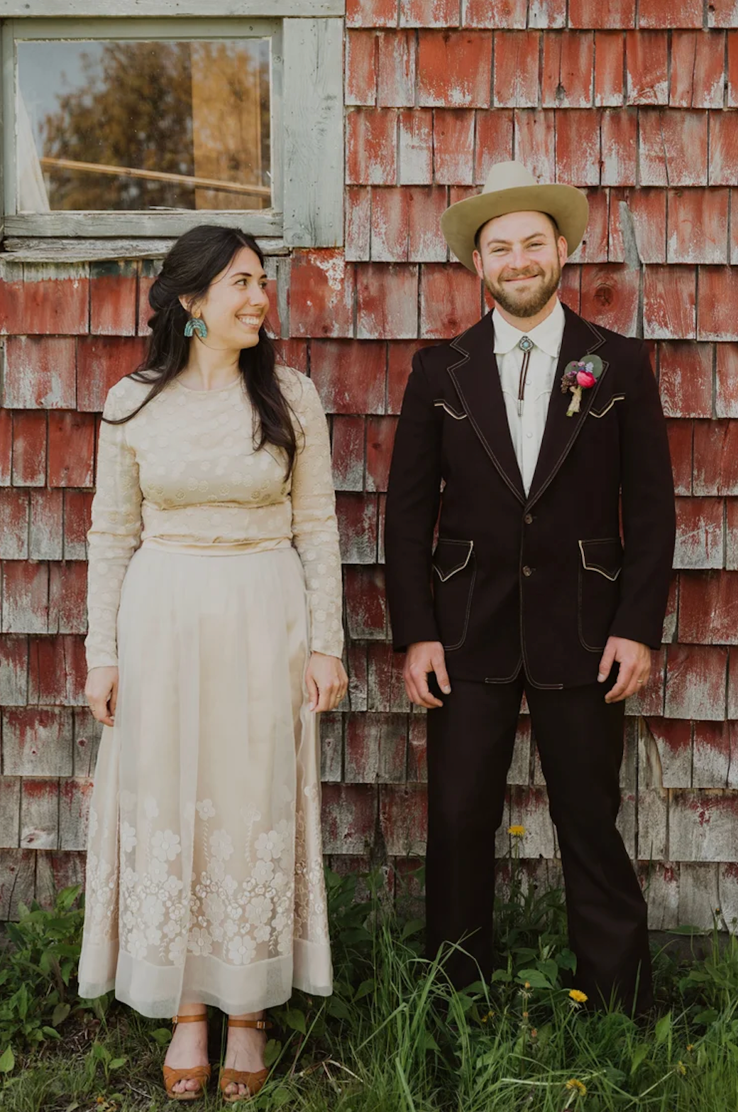 A bride in a vintage cream lace dress and groom in a dark suit with boutonniere pose before a rustic red barn.