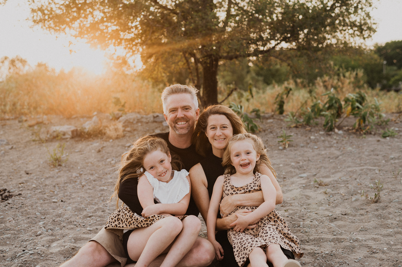 Happy family of four sitting together outdoors at golden hour, parents with two young daughters in matching leopard print skirts.