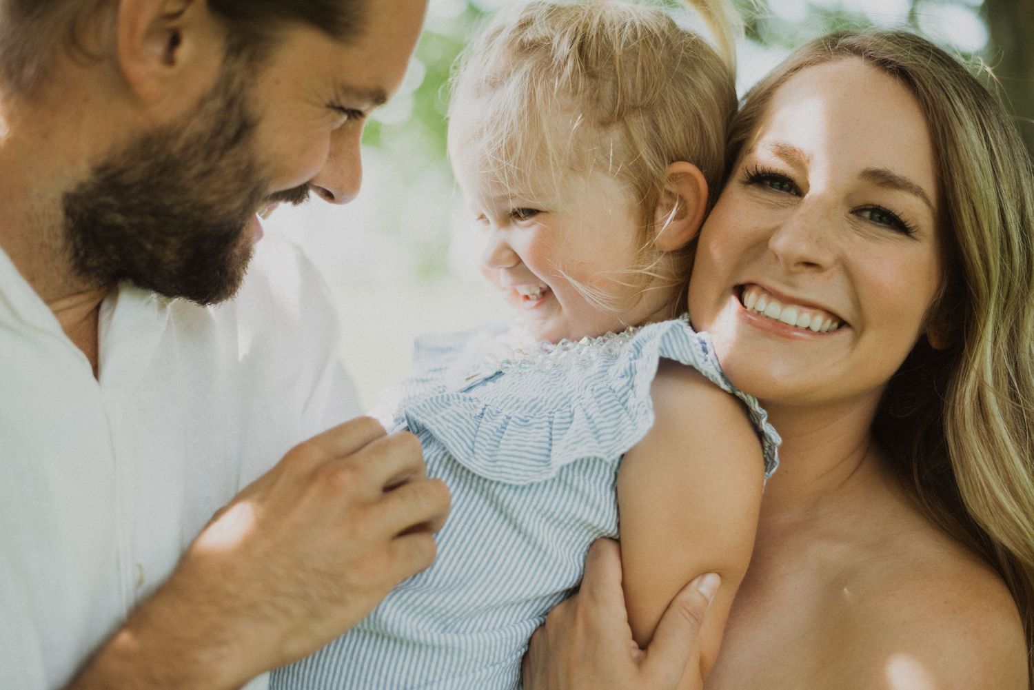 Happy family portrait with smiling parents holding their laughing toddler girl in a light blue dress outdoors.