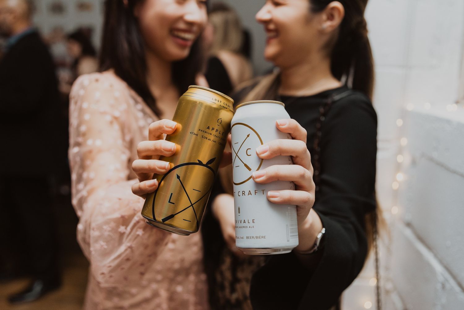 Two women clinking canned drinks together at a party, wearing elegant attire in a festive indoor setting.