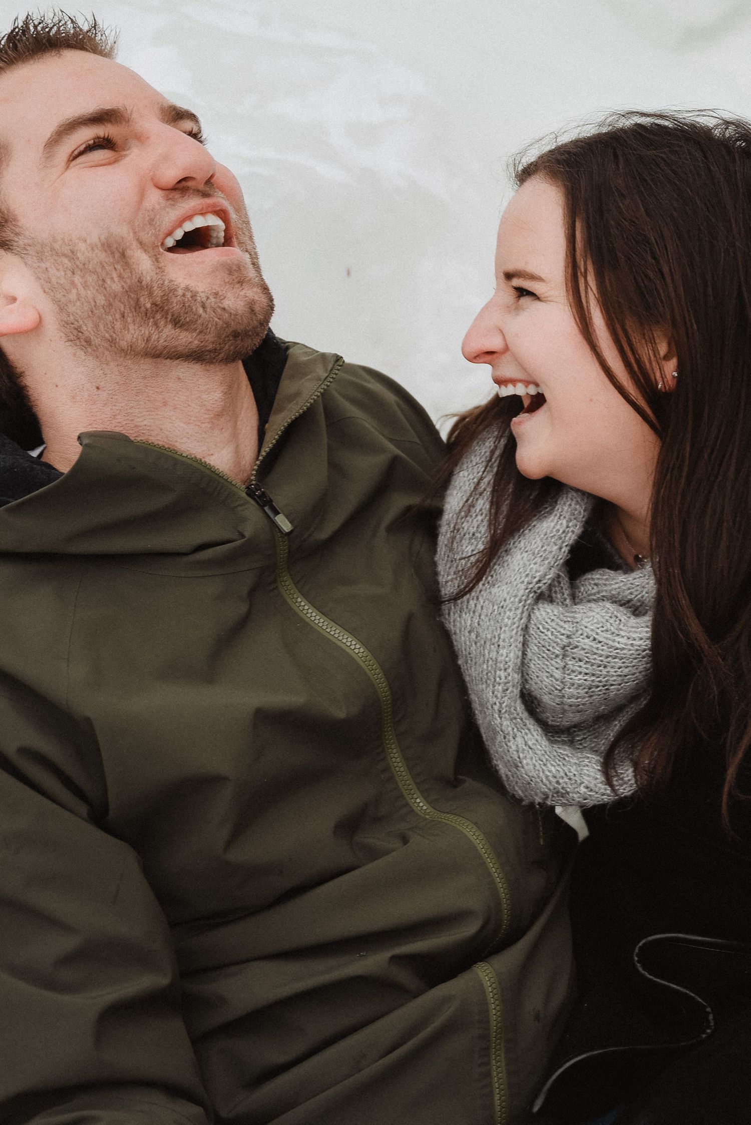 Happy couple laughing together outdoors in winter, woman wearing gray knit scarf, both in green jackets.