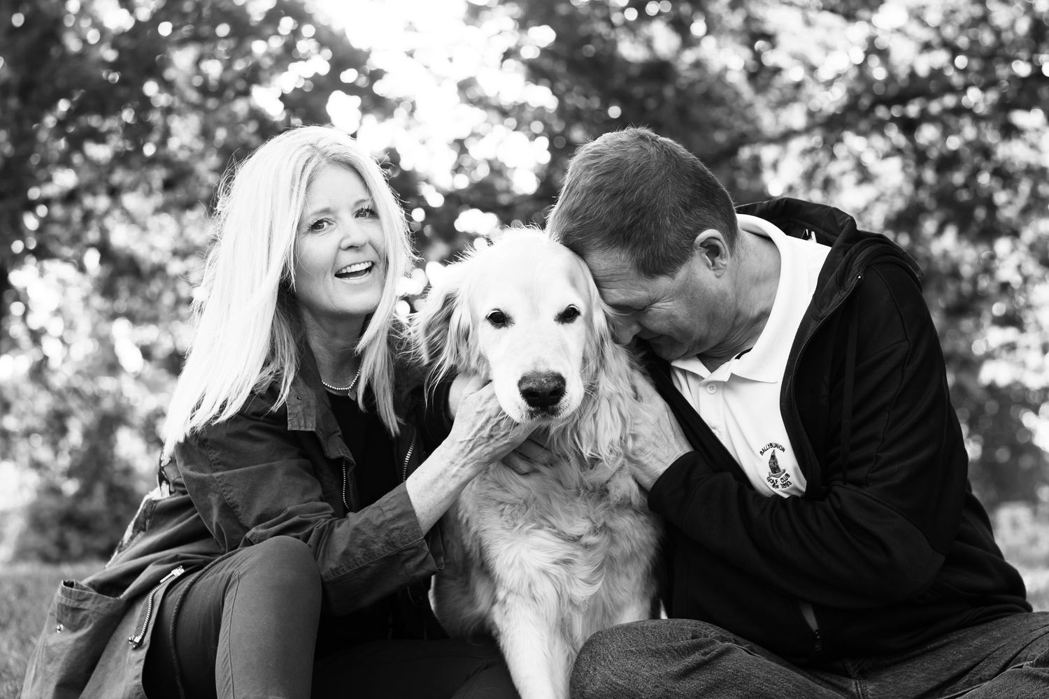 Black and white photo of a smiling couple with a golden retriever dog outdoors among trees.