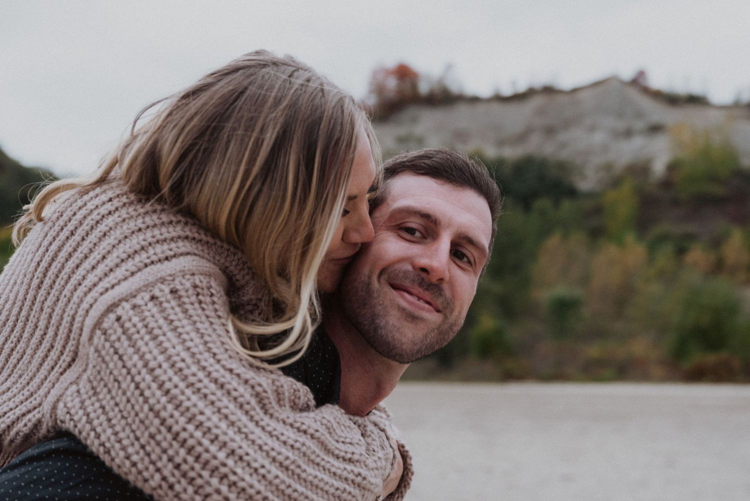 A smiling man gives a woman a piggyback ride outdoors, she leans in to kiss his cheek near a rocky landscape.