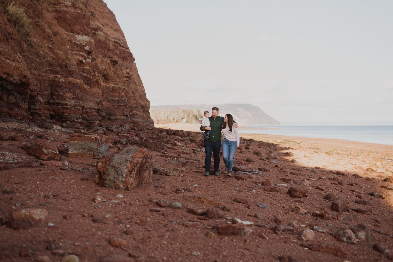 Couple standing on rocky red beach beside towering cliffs with calm ocean and distant headland in background.