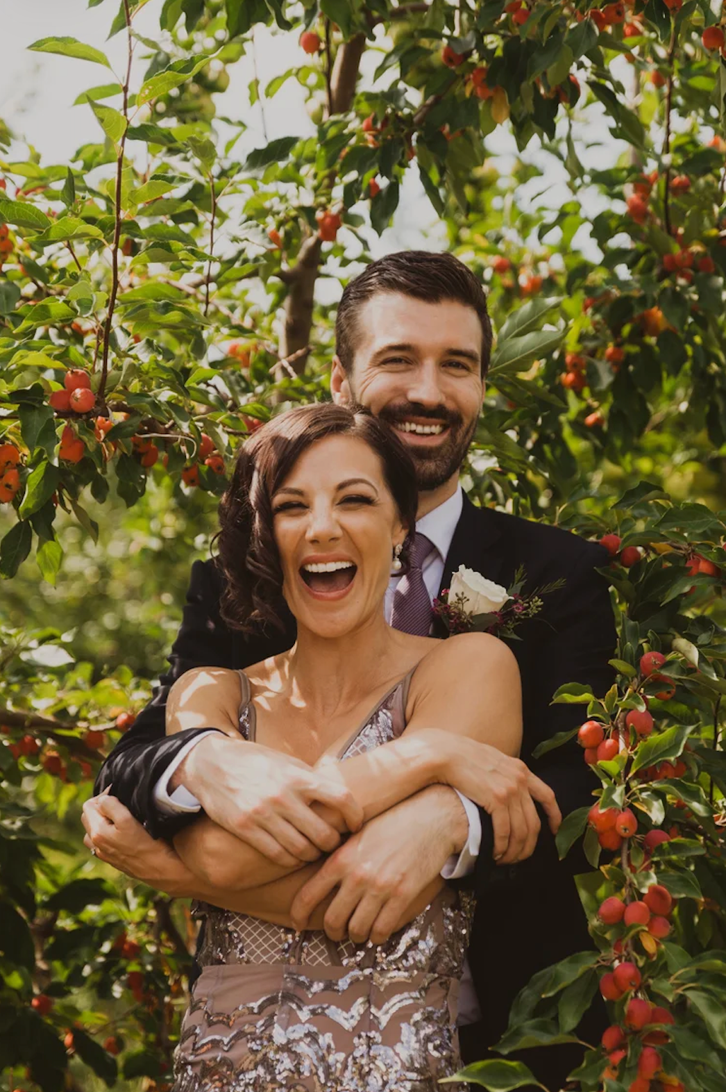 Happy couple embracing among cherry trees, bride in sequined dress and groom in suit with boutonniere.