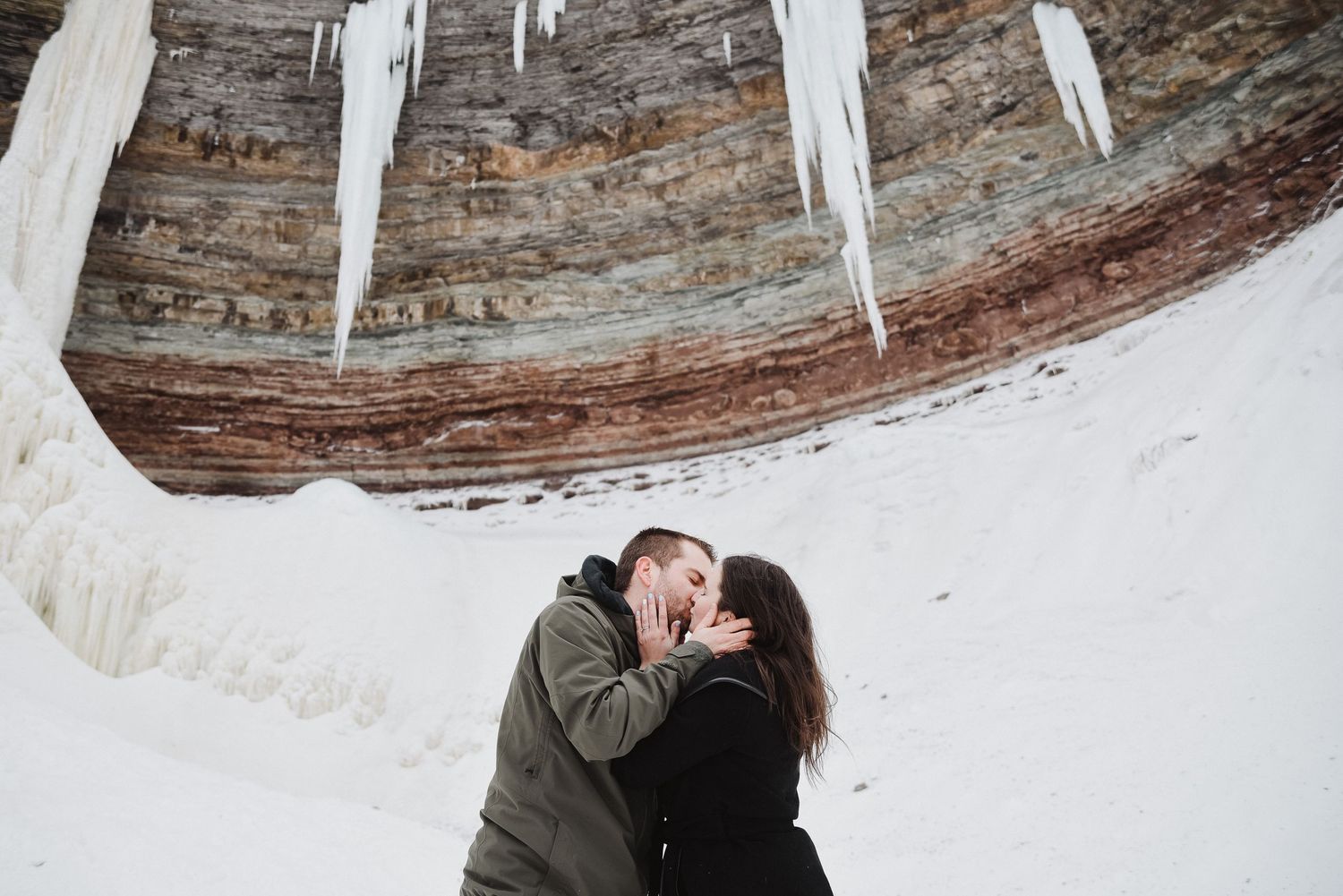 Couple sharing a romantic kiss under icy rock formation in snowy winter landscape.