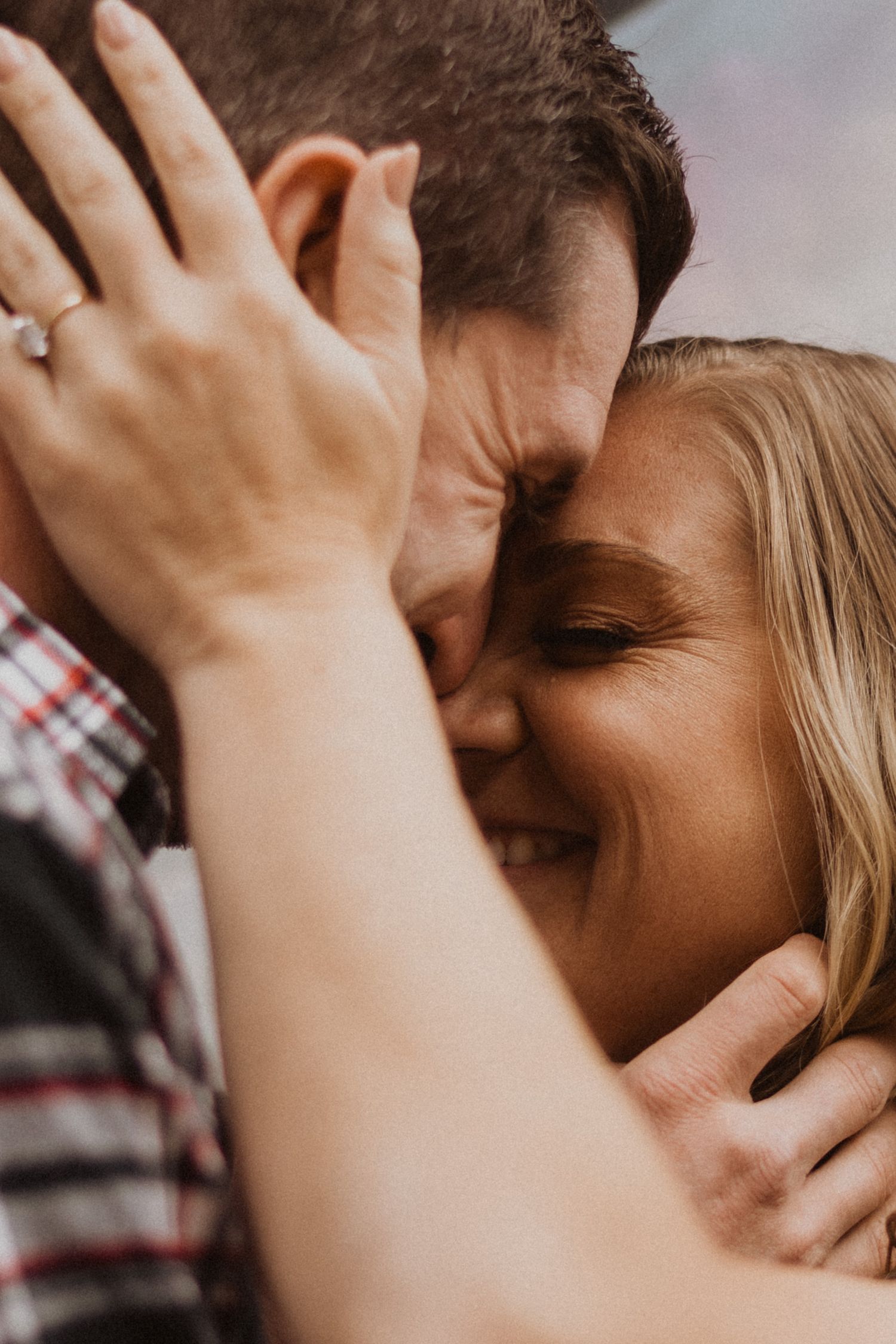 A joyful couple embraces closely, the woman smiling with an engagement ring visible on her hand.