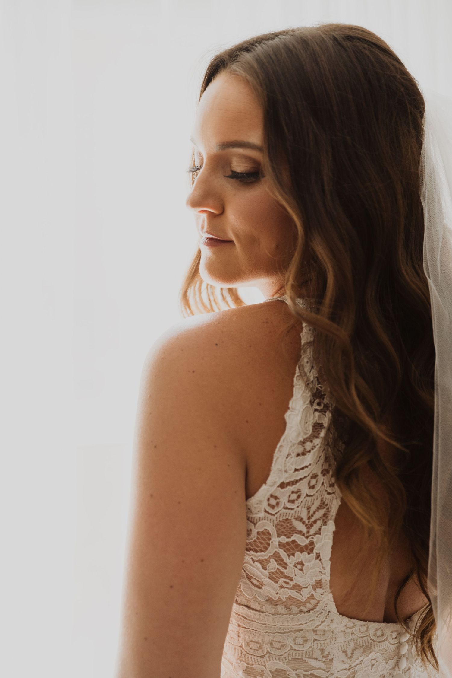 Bride in lace wedding dress with flowing brown hair, elegant makeup, looking down in soft natural light.