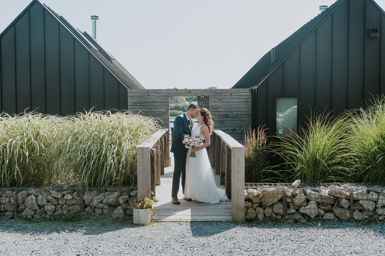 Bride and groom share a romantic moment on a wooden bridge surrounded by stone walls and ornamental grasses at a barn venue.