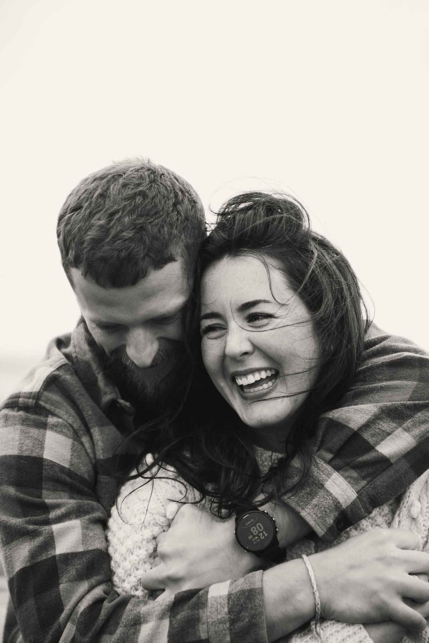 Black and white photo of a laughing woman being embraced from behind by a bearded man, both wearing plaid shirts.