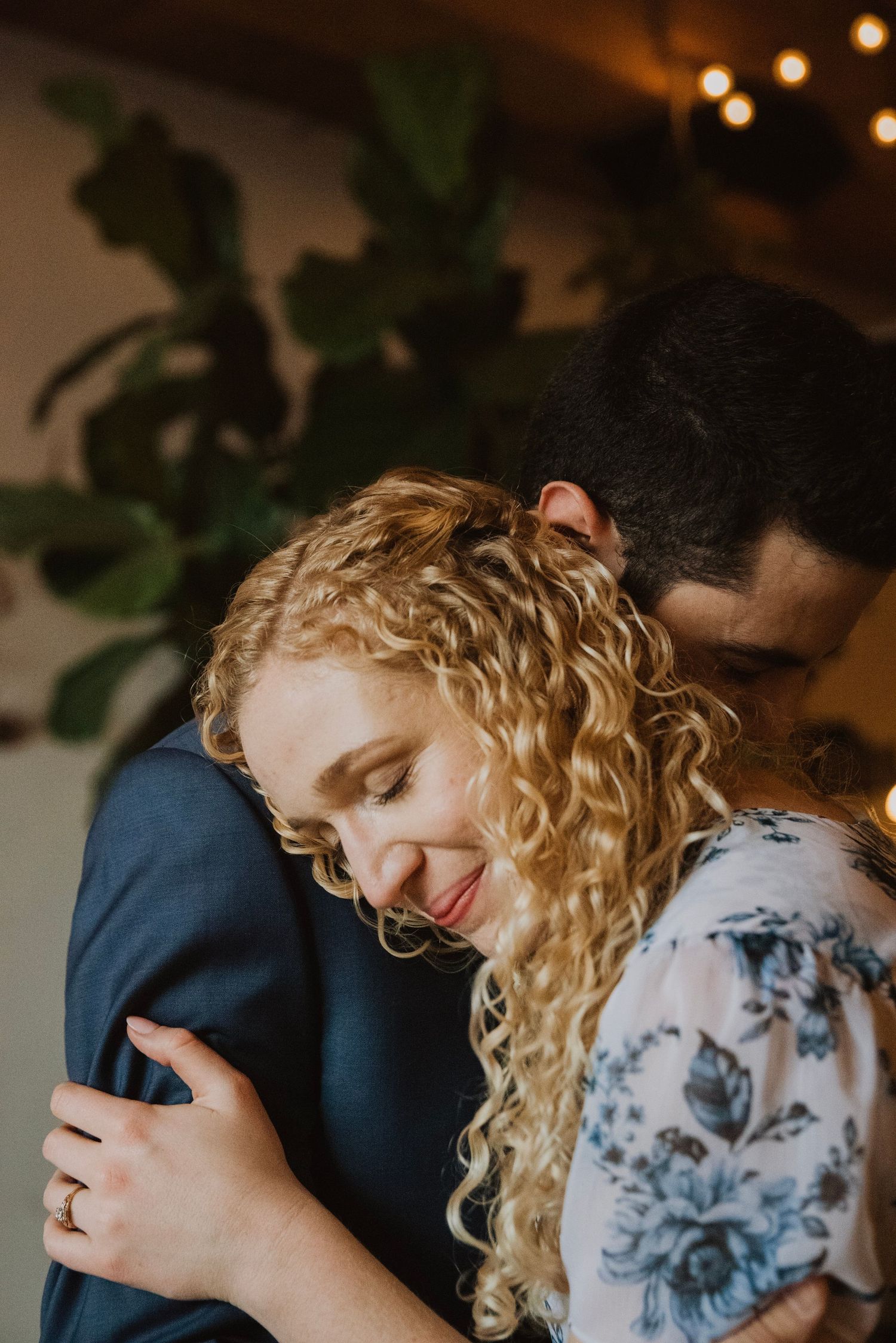 A woman with curly blonde hair smiles while embracing a man in a navy suit, with bokeh lights and greenery in the background.
