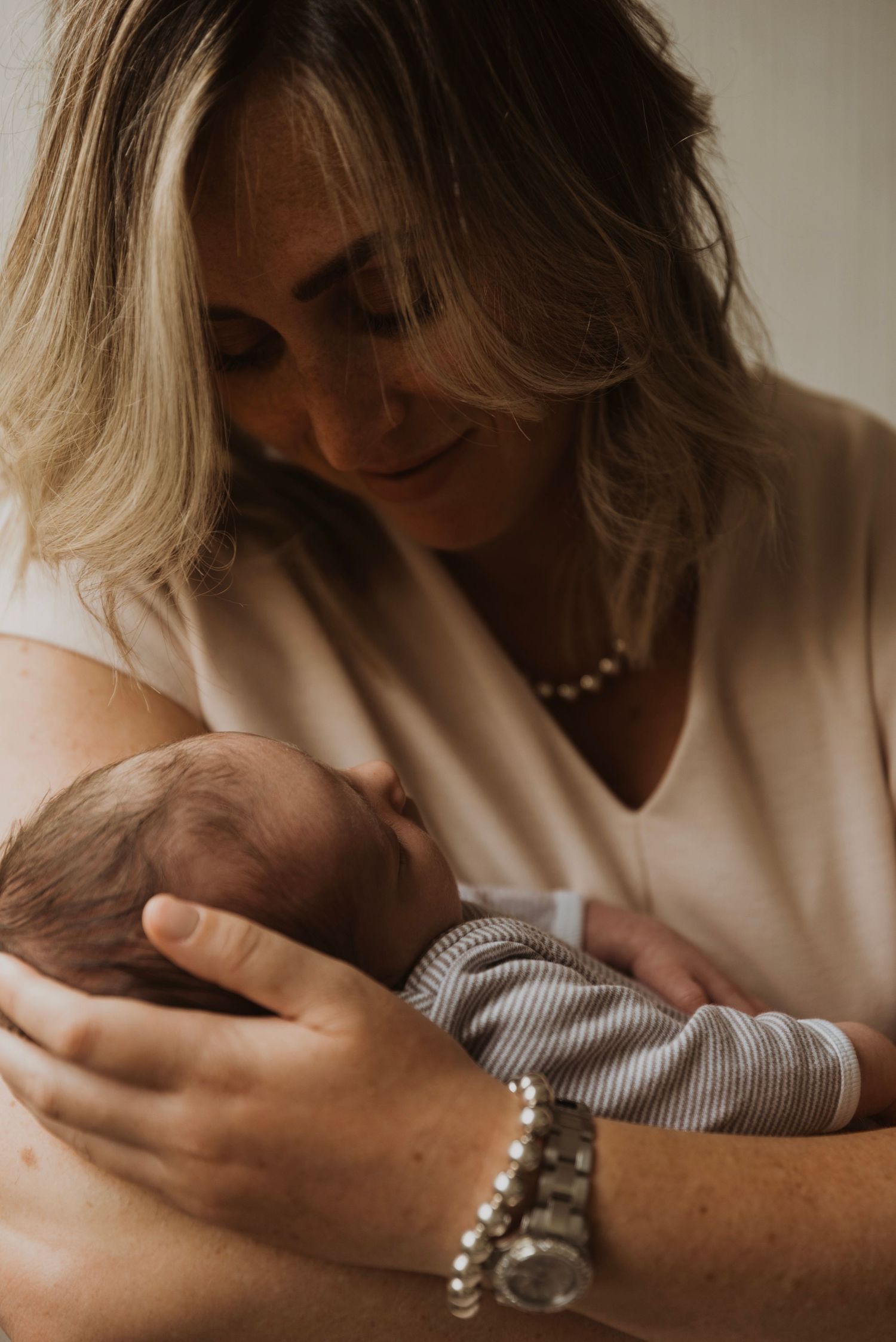 A smiling mother tenderly cradles her newborn baby, wearing a pearl necklace and silver bracelet.