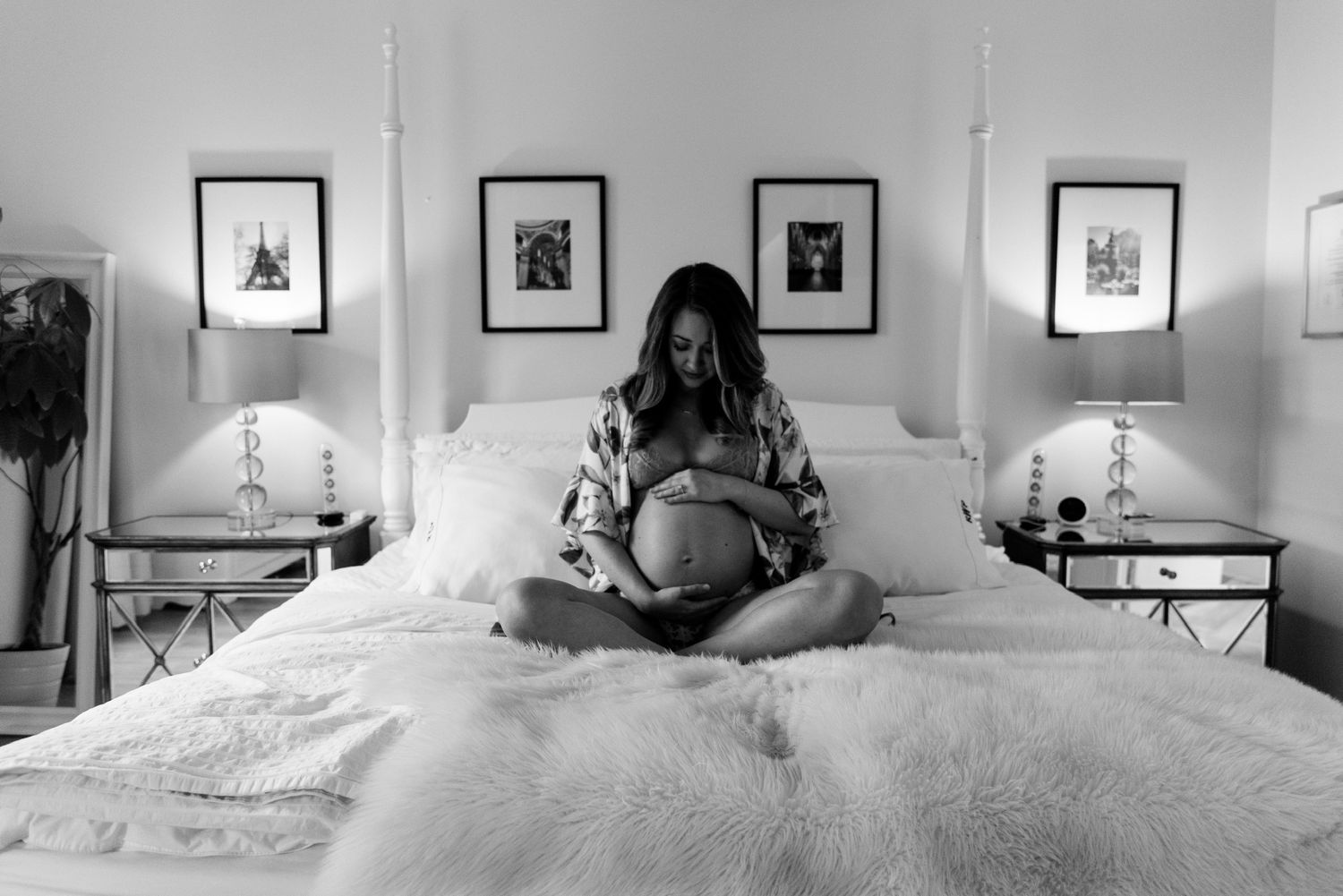 Black and white maternity photo of pregnant woman sitting cross-legged on bed cradling baby bump in serene bedroom.