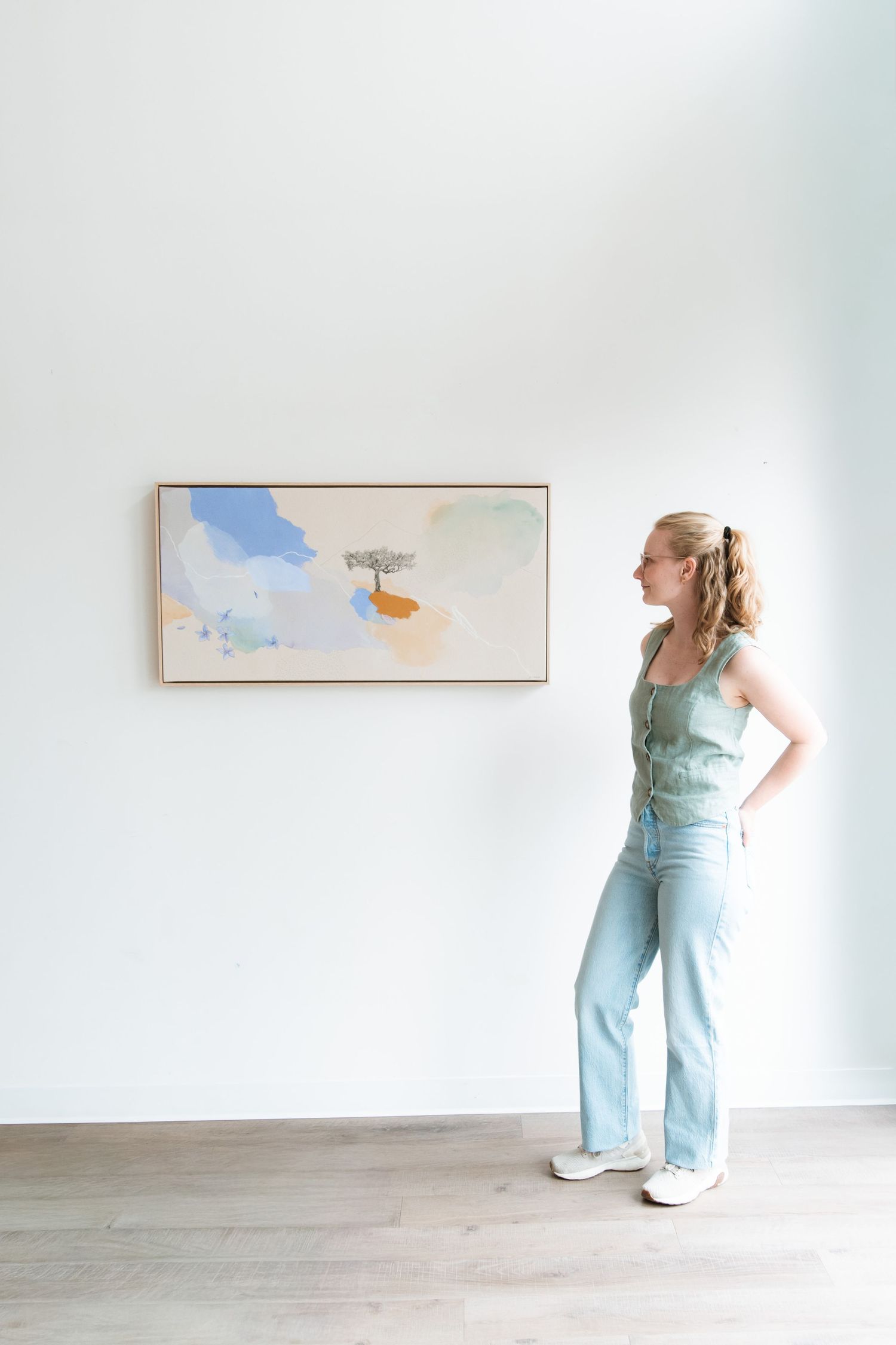 Woman in jeans and green top viewing abstract pastel painting hung on white gallery wall.