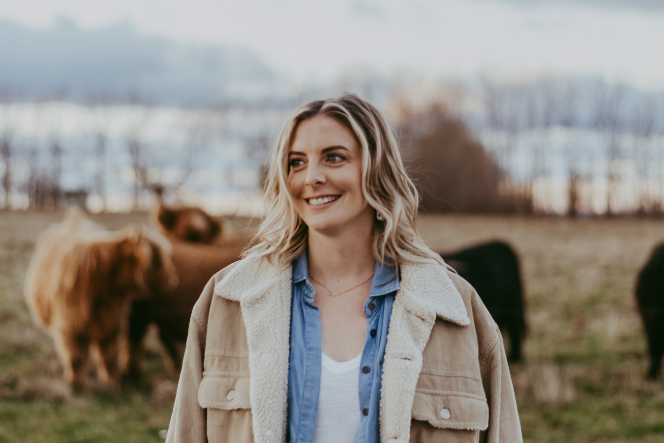 Smiling blonde woman in a beige shearling jacket standing in a field with Highland cattle in the background.