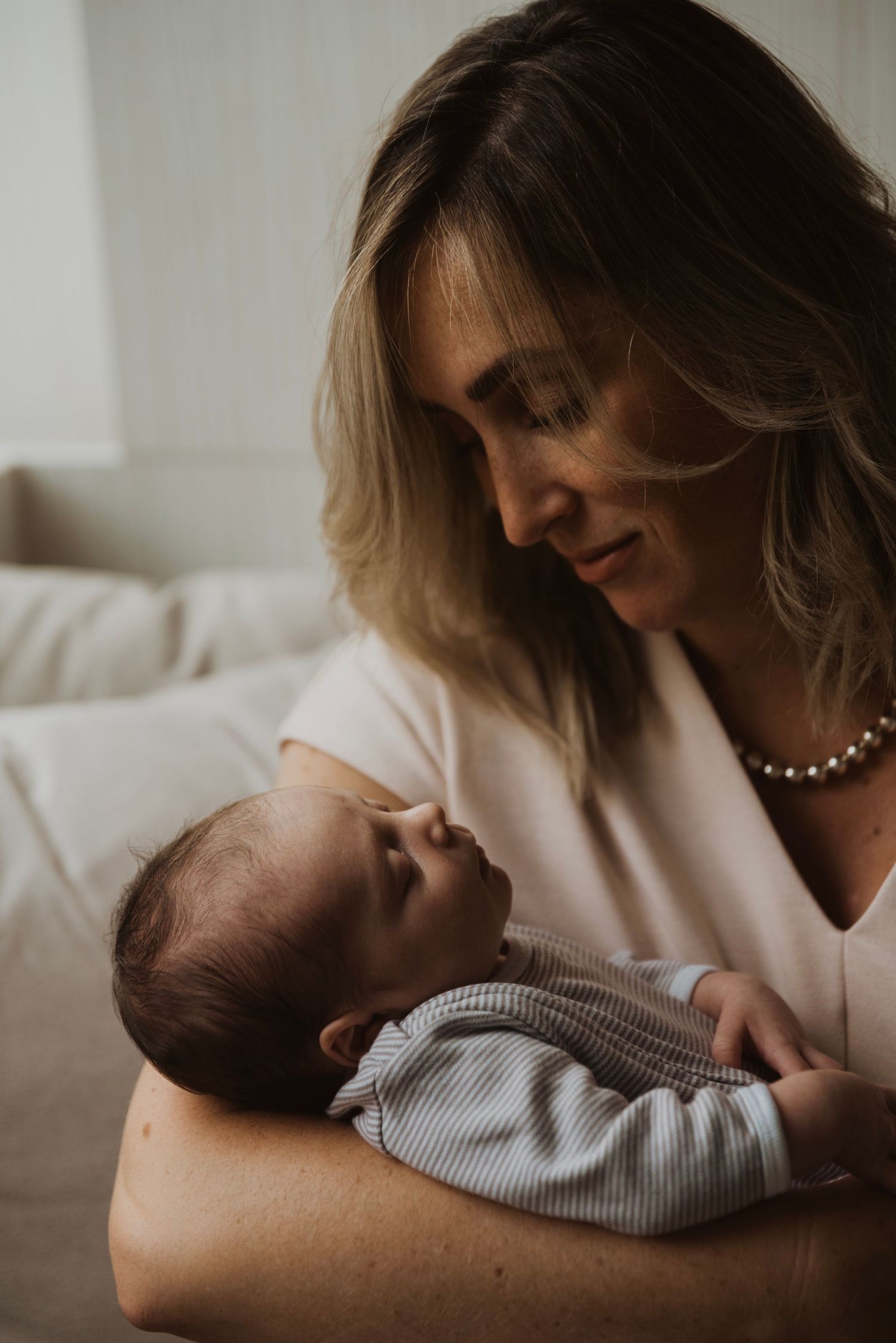 A mother tenderly gazes at her newborn baby cradled in her arms, wearing a pearl necklace.