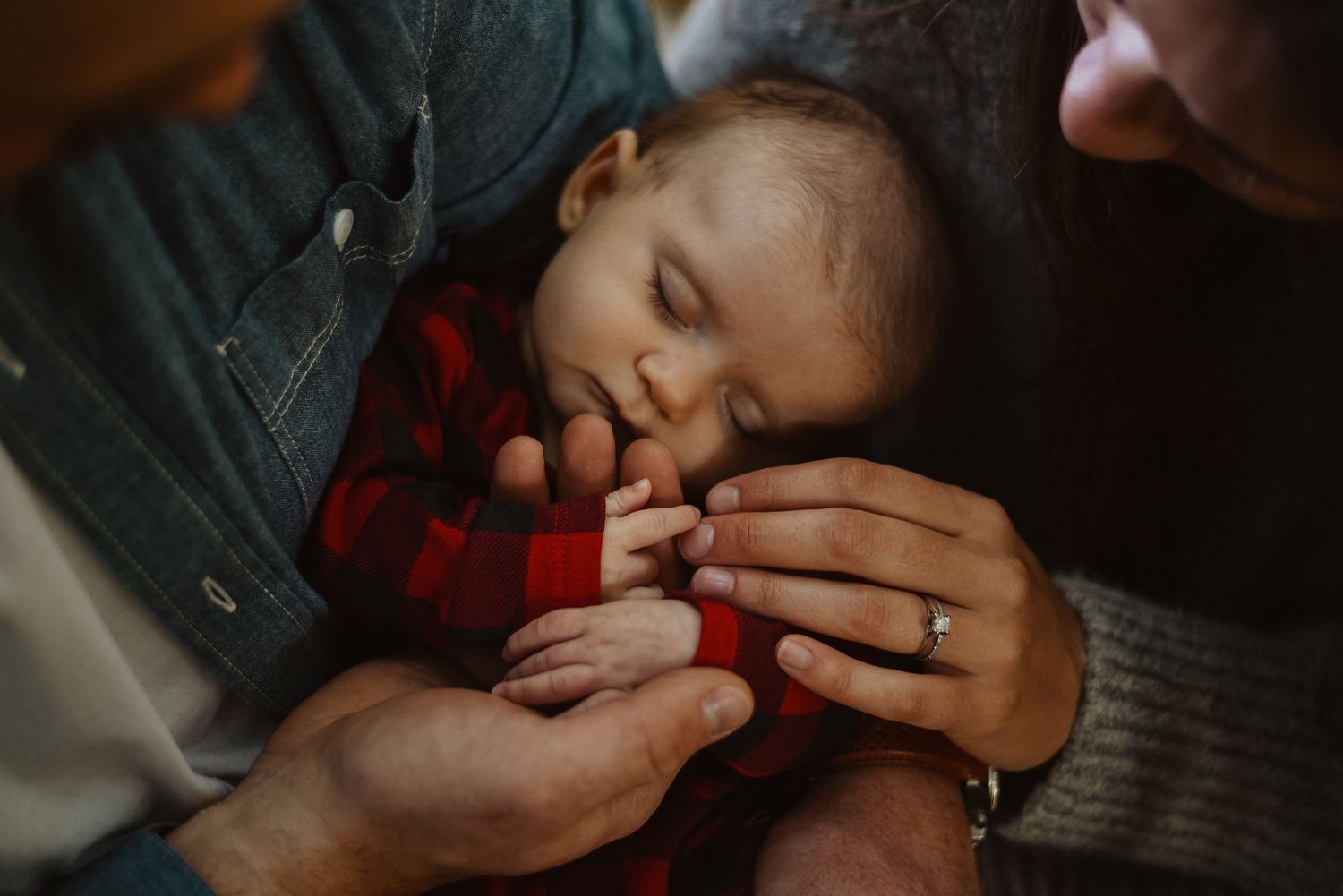 Sleeping newborn baby cradled in parents hands, wearing red plaid onesie, mother wearing wedding ring.