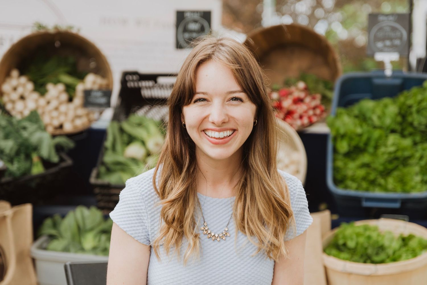 Smiling woman with long hair at a farmers market surrounded by fresh vegetables and greens.