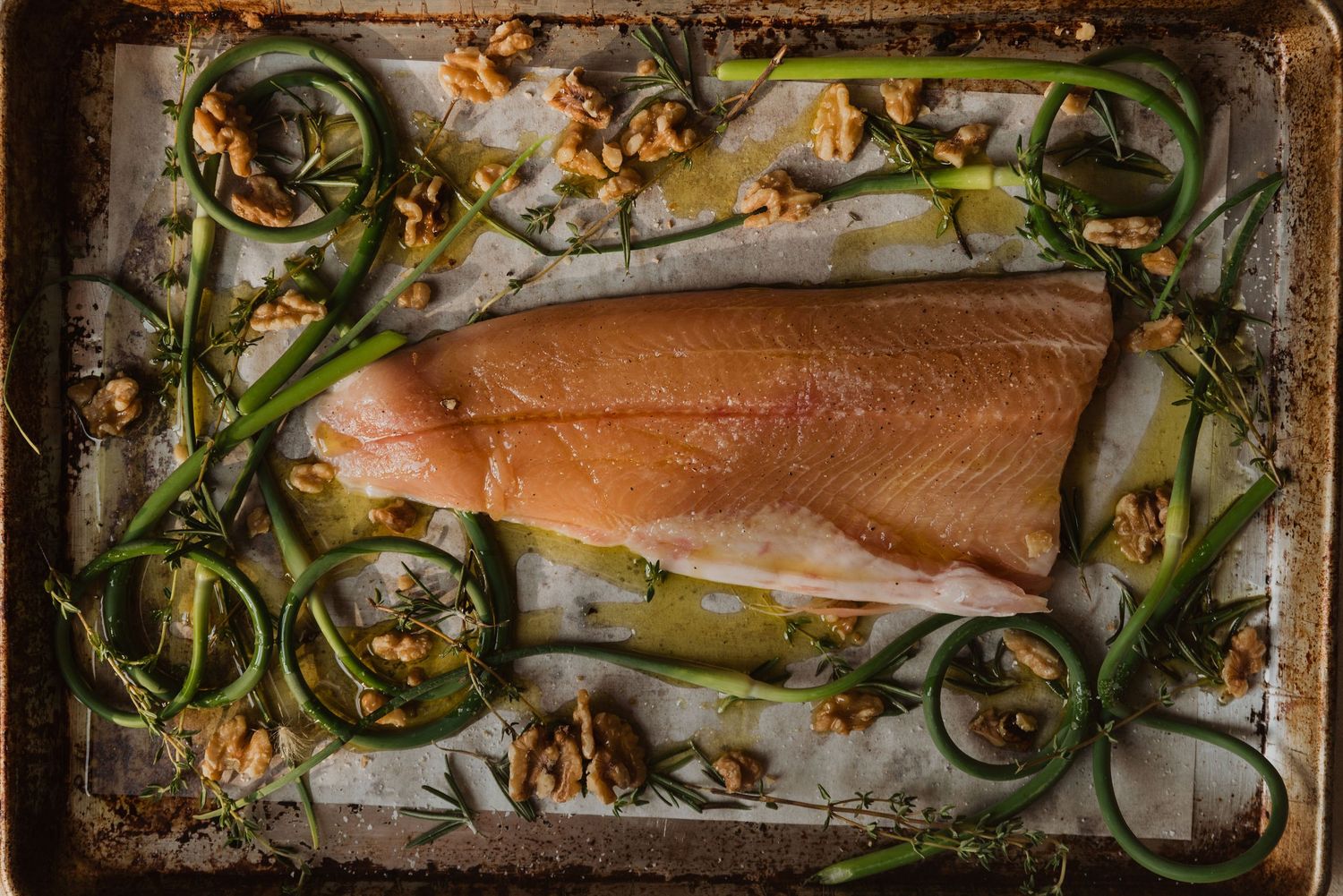 Raw salmon fillet on baking sheet surrounded by green onions, walnuts, and fresh herbs ready for roasting.