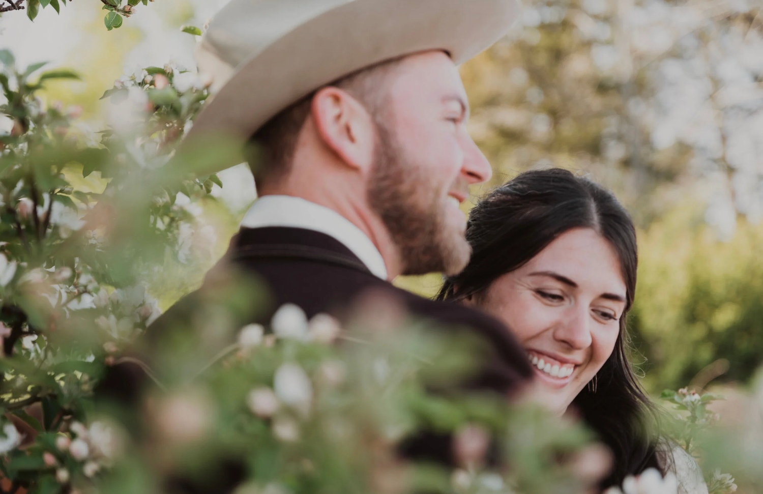 Couple smiling together outdoors among blooming flowers, man wearing white cowboy hat, romantic countryside wedding photo.