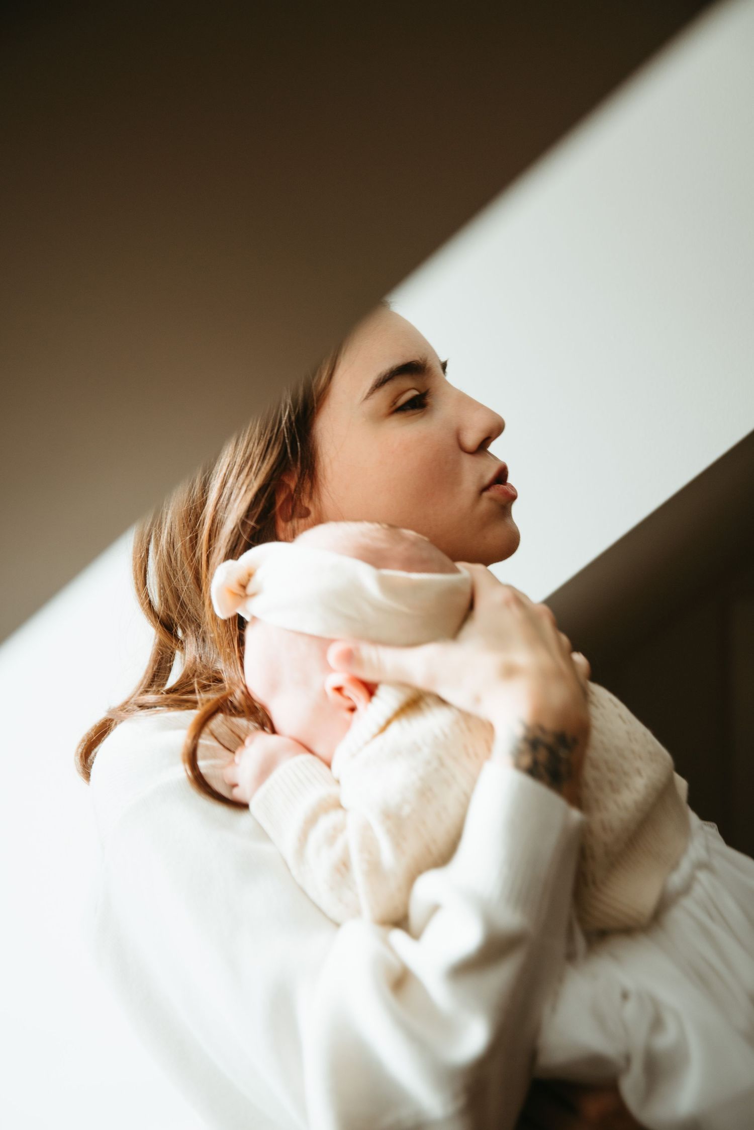 A young mother tenderly holds her newborn baby against her shoulder in soft natural light.