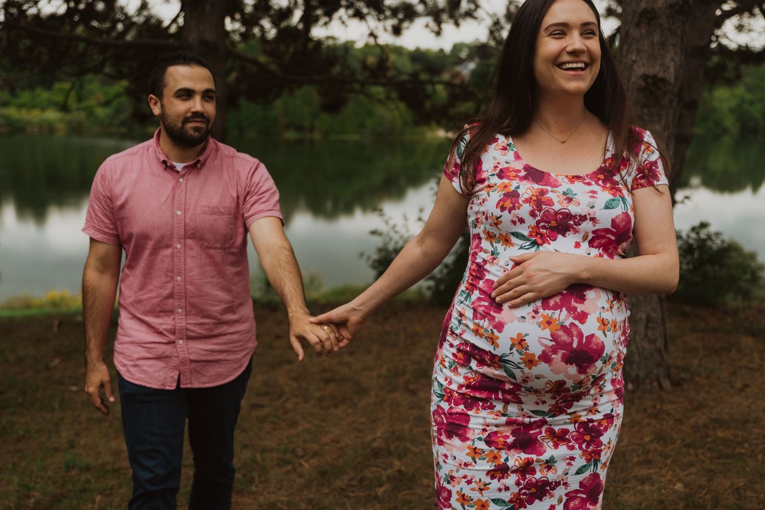 Pregnant woman in floral dress holds hands with man in pink shirt outdoors near a lake.
