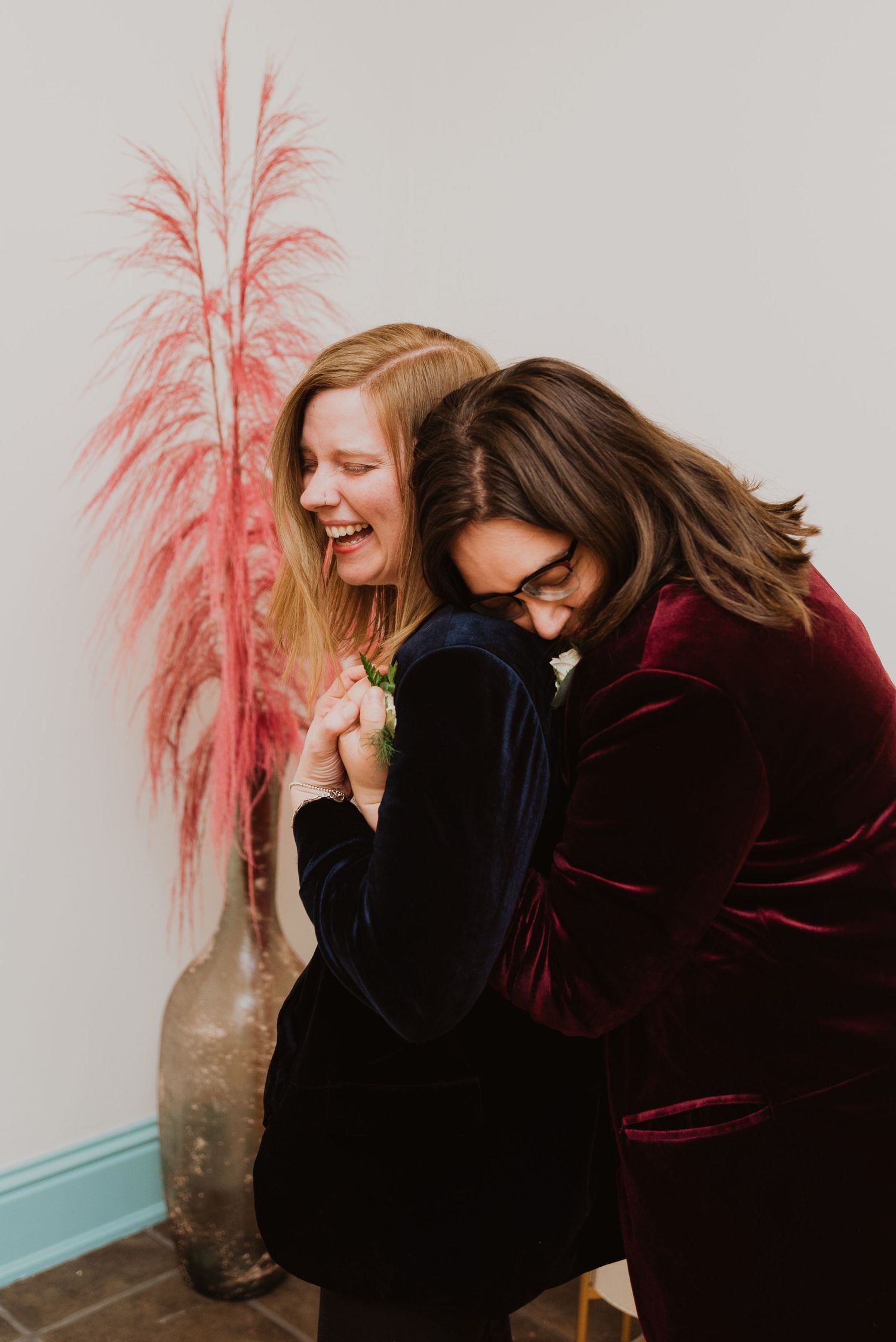Two women in velvet blazers laughing and embracing near a pink pampas grass arrangement at a celebration.
