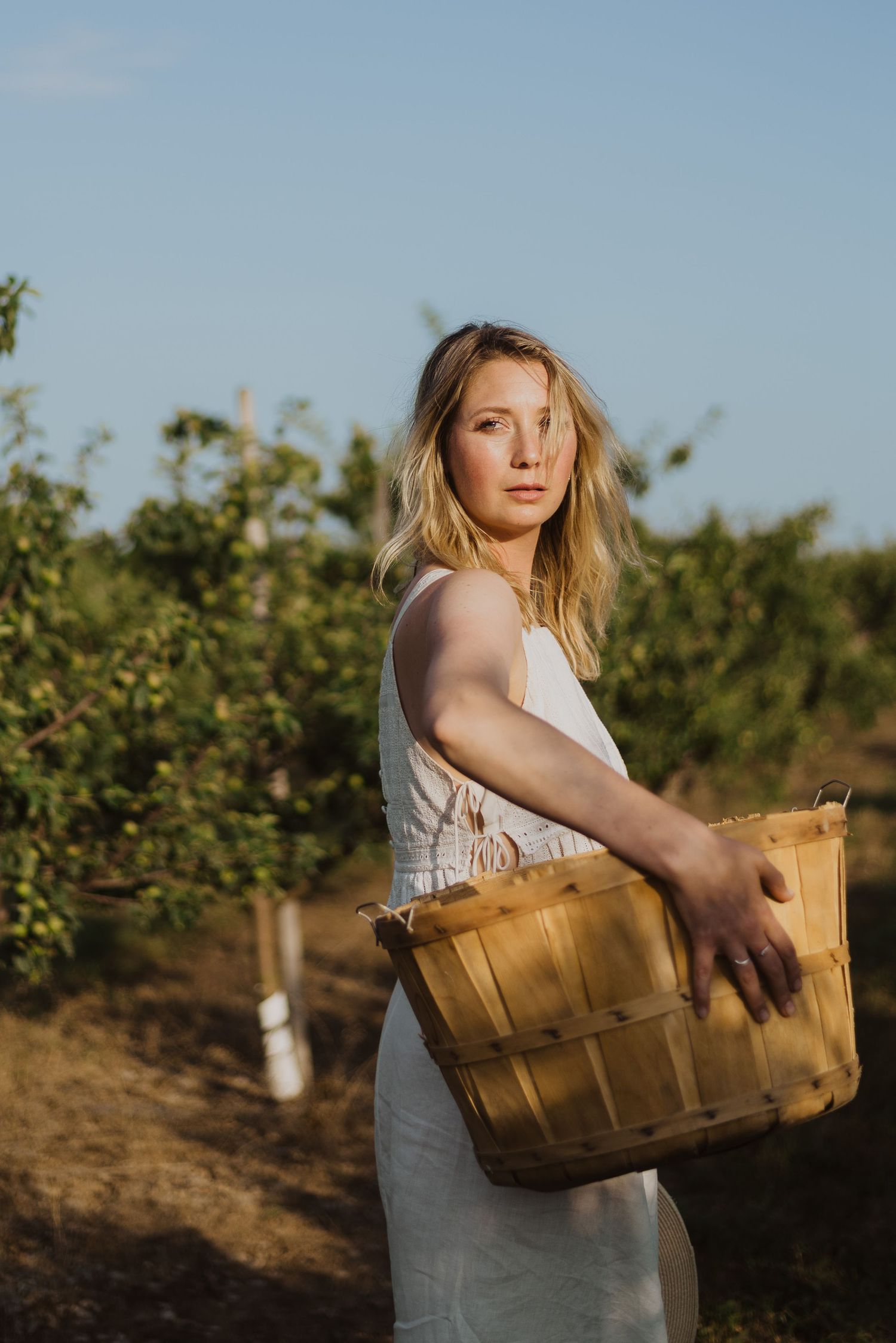A blonde woman in a white dress holds a wooden basket while standing in a lush orchard during golden hour.
